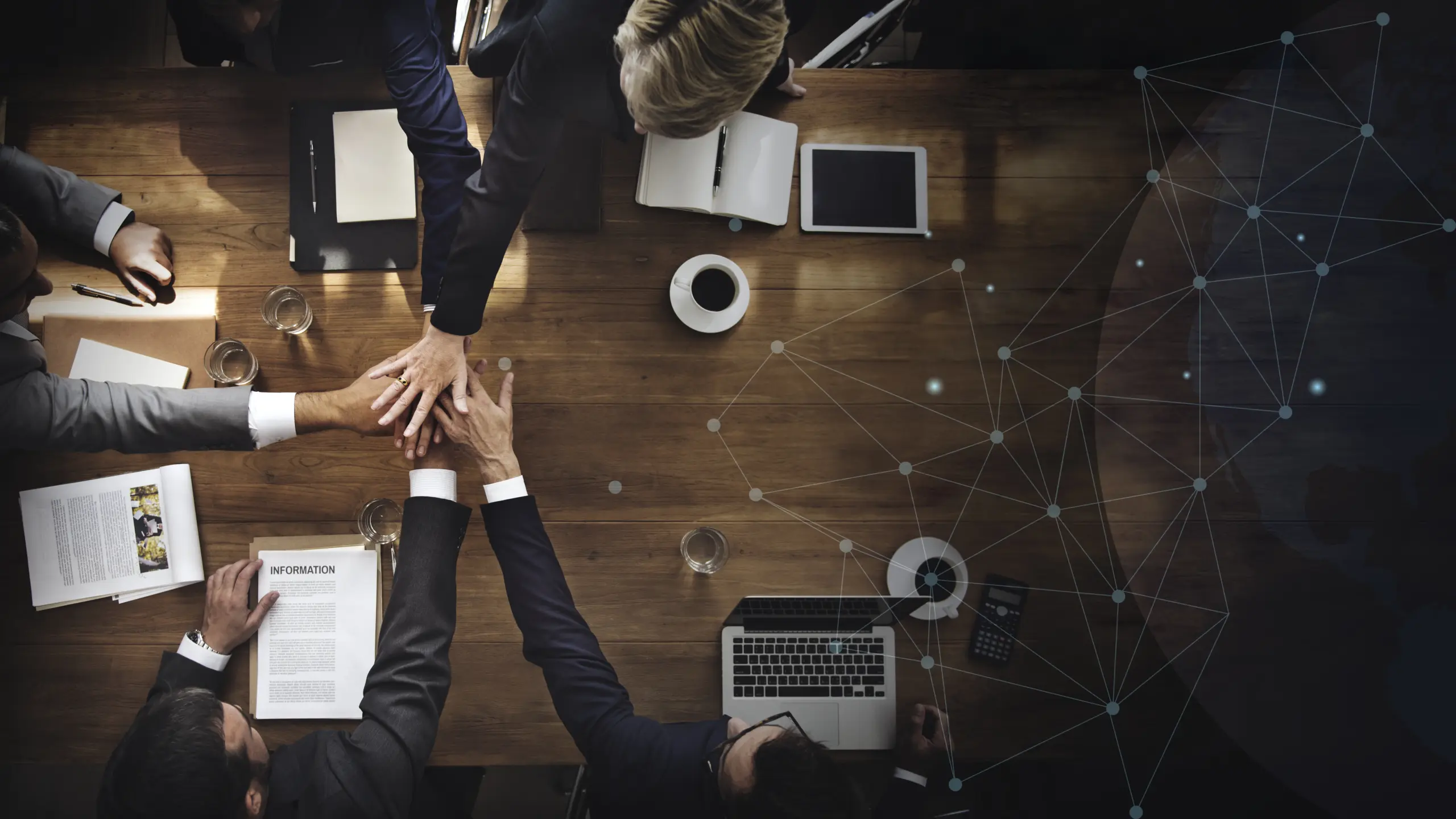 Mehrere Personen sitzen um einen Holztisch mit Laptops, Notizbüchern und Kaffeetassen und legen ihre Hände in der Mitte zusammen, als Symbol für Teamwork und digitale Zusammenarbeit. Rechts ist ein digitales Netzwerk-Overlay zu sehen, das Konnektivität und Technologieintegration darstellt.